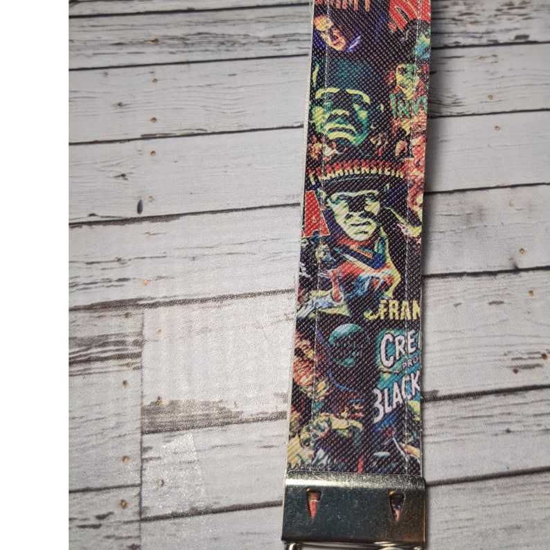 May include: A white fabric keychain with a colorful print of classic horror movie posters. The print features images of Frankenstein, Dracula, and other monsters. The keychain has a silver metal clasp.
