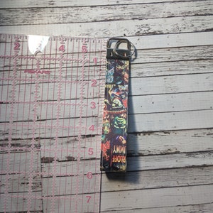 May include: A black and green key chain with a colorful graphic print of movie monsters. The key chain is approximately 6 inches long and has a silver metal key ring.