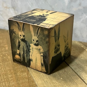May include: A wooden cube with a vintage image of three rabbits. The rabbits are dressed in clothing and have a vintage look. The cube is a light brown color and has a distressed finish.