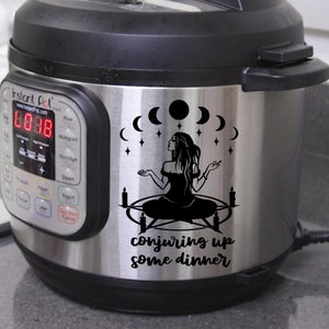 May include: A black and white sticker featuring a witch conjuring up some dinner. The sticker is applied to a silver Instant Pot with a digital display. The sticker depicts a witch with long hair, seated in a circle with candles and moons.