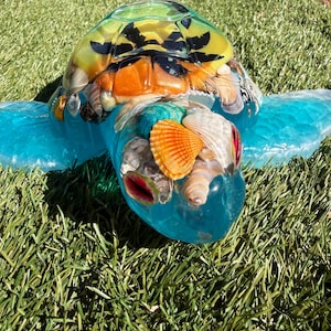 May include: A clear resin sea turtle figurine featuring a translucent blue body and a shell filled with various embedded seashells in shades of orange, white, and brown, with black accents on the top of the shell and a bright orange shell on the head.