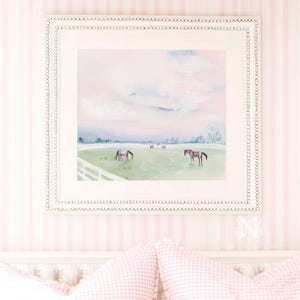 May include: A square framed painting featuring a landscape with horses grazing in a green pasture behind a white fence. The sky is painted in soft pastel pink and blue tones. The frame is white with a decorative scalloped edge detail.