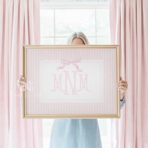 Op de afbeelding: Een rechthoekige goudkleurige lijst met een print op een lichtroze en wit verticaal gestreepte achtergrond. De print bevat een gecentreerde witte rechthoekige rand, een roze aquarel strik bovenaan en de roze monogramletters M, N en M.