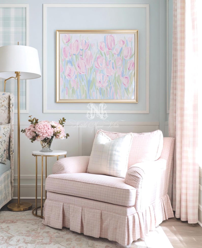 May include: A framed painting of pink tulips in a gold frame hangs above a light pink upholstered armchair with a pleated skirt. The chair features a white plaid throw pillow. A gold floor lamp and a small table with pink flowers are nearby.