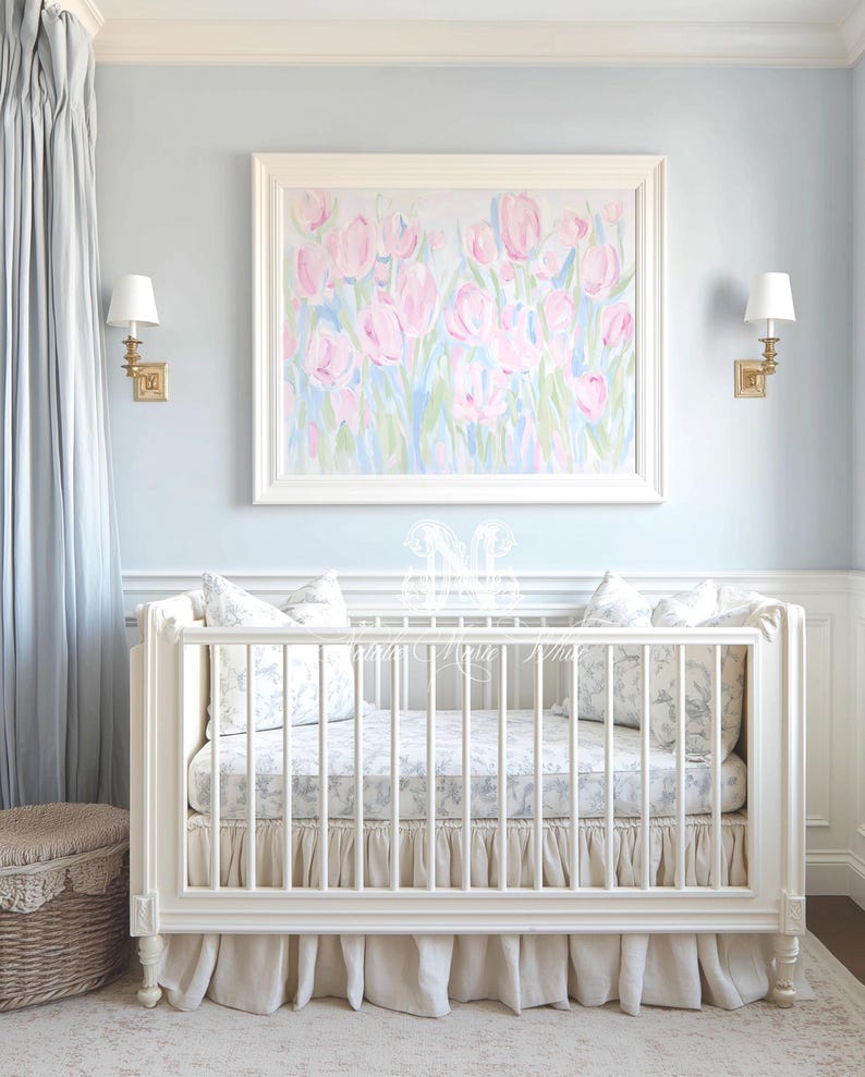 May include: A white wooden crib with vertical slats, a ruffled beige bed skirt, and a mattress featuring a grey and white floral pattern. Two matching floral pillows sit inside the crib, positioned beneath a framed painting of pink tulips.