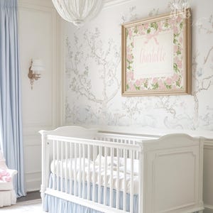 May include: A white wooden cot with a light blue ruffled skirt, positioned beneath a framed rectangular wall art piece featuring a pink floral border, a pink bow, and the name Charlotte printed in pink script on a white background.
