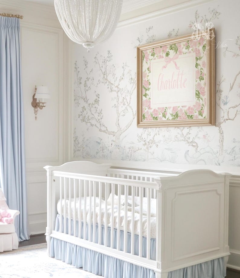 May include: A white wooden cot with a light blue ruffled skirt, positioned beneath a framed rectangular wall art piece featuring a pink floral border, a pink bow, and the name Charlotte printed in pink script on a white background.