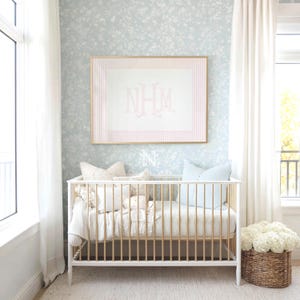 May include: A white and gold crib with vertical slats, featuring a light blue floral wallpaper background. Above the crib hangs a framed print with pink vertical stripes and the monogram NHM in the center. A woven basket with white flowers sits nearby.