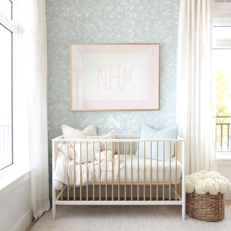 May include: A white and gold crib with vertical slats, featuring a light blue floral wallpaper background. Above the crib hangs a framed print with pink vertical stripes and the monogram NHM in the center. A woven basket with white flowers sits nearby.