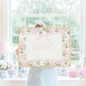 May include: A rectangular print featuring a light pink floral hydrangea pattern surrounding a central white rectangular border. A light pink ribbon bow is centred at the top of the border, which contains the name Charlotte in pink script.