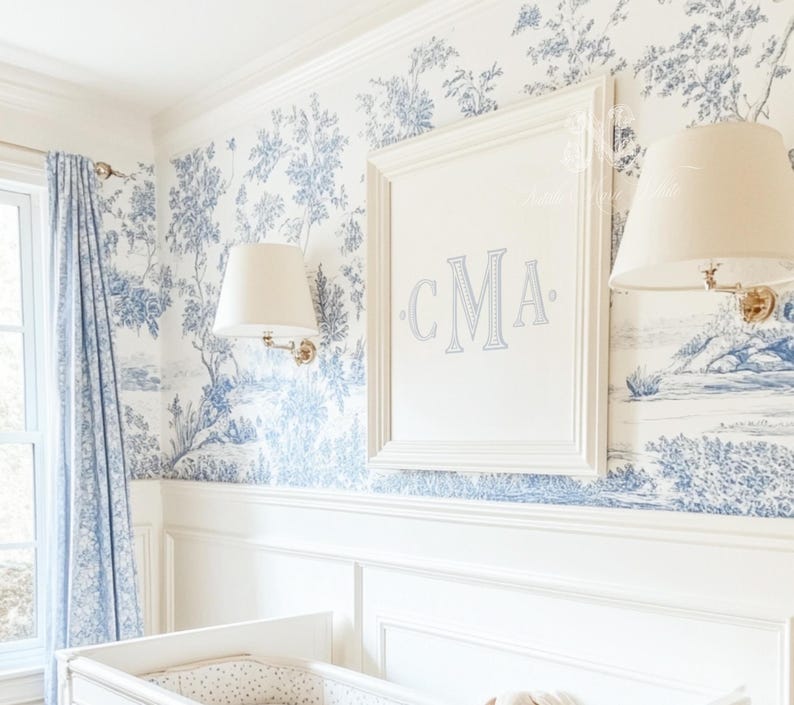 May include: A white framed picture with the initials "CMA" in blue embroidery on a white background. The picture is hanging on a wall with a blue and white floral wallpaper pattern.
