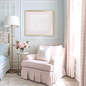 May include: A light pink upholstered armchair with a pleated skirt and a white plaid throw pillow. Beside it, a gold metal side table holds a glass vase with pink hydrangeas. Above, a gold-framed print displays the monogram NHM in pink.