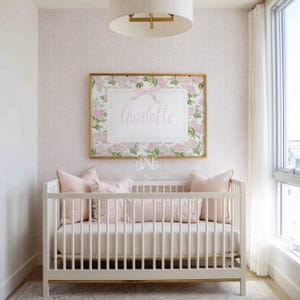 May include: A white wooden cot with a cream mattress and three light pink pillows, positioned beneath a rectangular wall art piece featuring a floral border, a pink ribbon graphic, and the name Charlotte in elegant script lettering.