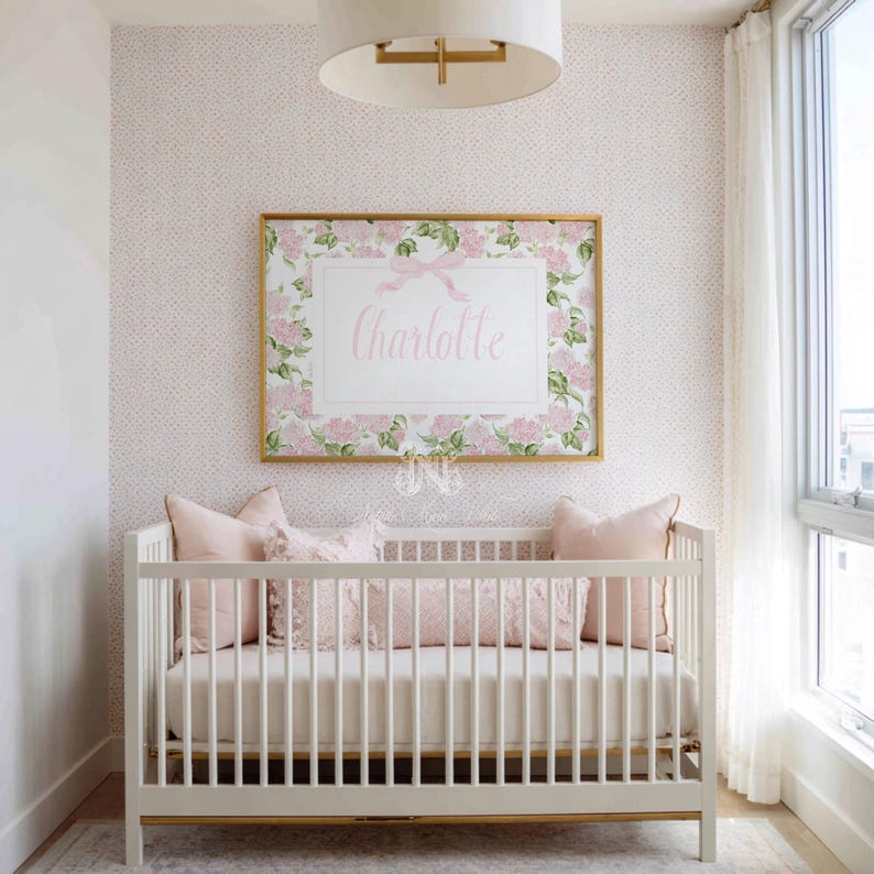 May include: A white wooden cot with a cream mattress and three light pink pillows, positioned beneath a rectangular wall art piece featuring a floral border, a pink ribbon graphic, and the name Charlotte in elegant script lettering.