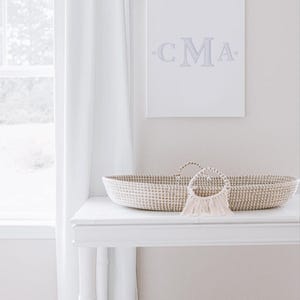 May include: A white wicker bassinet with a white and beige woven design, a white table with a white top and bamboo legs, and a white canvas with the text "CMA" in gray lettering.