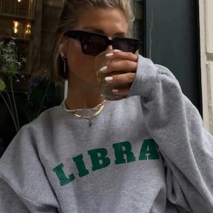 May include: A grey sweatshirt with the word "LIBRA" printed in green letters. The person wearing the sweatshirt is holding a glass of liquid and wearing black sunglasses.