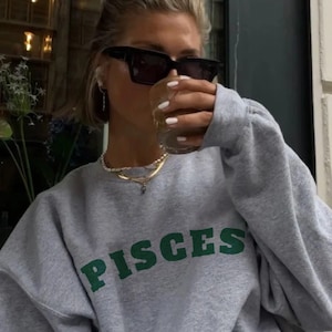 May include: A woman wearing a gray sweatshirt with the word "PISCES" printed in green letters. She is wearing black sunglasses and holding a clear glass with a light-colored beverage.