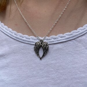 Puede incluir: Collar plateado con un colgante en forma de alas de &aacute;ngel. Las alas, con detalles finos, cuelgan de una cadena delicada. El collar se muestra sobre una camiseta blanca, creando un estilo sencillo.