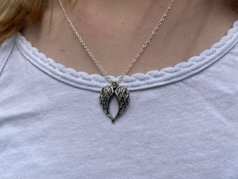 Puede incluir: Collar plateado con un colgante en forma de alas de &aacute;ngel. Las alas, con detalles finos, cuelgan de una cadena delicada. El collar se muestra sobre una camiseta blanca, creando un estilo sencillo.