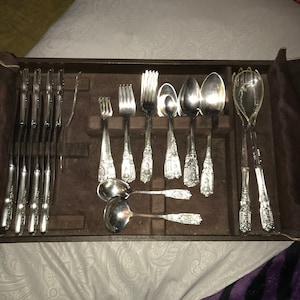 Puede incluir: Un juego de cubiertos de plata en un estuche de cuero marrón. El juego incluye tenedores, cuchillos, cucharas y una cuchara para servir.