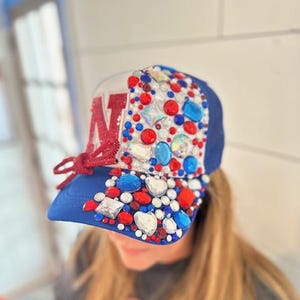 May include: A blue and white trucker hat with a red letter "N" on the front. The hat is decorated with rhinestones in red, white, and blue.