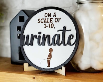 May include: A round wooden sign with a black border and white background. The sign says "On a scale of 1-10, urinate" in black lettering. A small silhouette of a person urinating is below the text. The sign is on a small wooden stand.