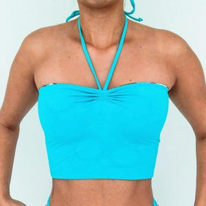 May include: A bright turquoise halter top with a gathered bust and a bandeau-style design. The top features a halter neck tie and a contrasting patterned lining visible at the top edge. This swimwear top is designed for a comfortable fit.