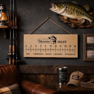 May include: A rectangular wooden wall sign labelled Fisherman's Ruler with a 20-inch measurement scale, fish graphics, and text categories including Bait, Throw Back, Keeper, Fish Story, and Outright Lie, suspended by a brown twine rope.