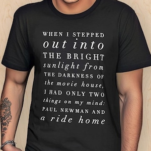 May include: A black short-sleeved t-shirt featuring white text that reads: WHEN I STEPPED out into THE BRIGHT sunlight from THE DARKNESS of the movie house, I HAD ONLY two things on my mind: PAUL NEWMAN AND a ride home.
