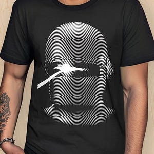 May include: Black t-shirt featuring a white graphic of a stylized face with a visor and a cigarette. The shirt has a crew neck and short sleeves. The text "THE MIDNIGHT LAUNDRY" is printed vertically on the left side.