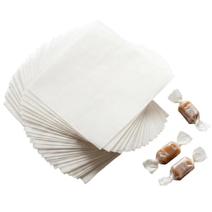 May include: A stack of white, square parchment paper sheets next to three individually wrapped caramel candies. The candies are in clear cellophane wrappers with twisted ends, showcasing the caramel's rich brown color.