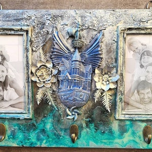 Könnte beinhalten: Ein rustikaler Holzbilderrahmen mit drei Fotoschlitzen, die jeweils ein Schwarzweißfoto einer Familie zeigen. Der Rahmen ist mit einem blauen und silbernen 3D-Verzierungsstück eines Hauses mit Flügeln verziert, das von Blumen umgeben ist.