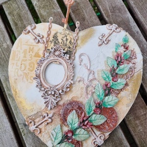 Puede incluir: Una placa de madera en forma de corazón decorada con una variedad de adornos, incluyendo una llave, una cadena, un marco pequeño y un diseño floral. La placa está pintada en tonos de blanco, dorado y marrón.