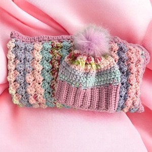 May include: A crocheted hat and scarf set in pastel colors of pink, blue, and lavender. The hat has a ribbed brim and a fluffy, light purple pom-pom. The scarf is rectangular with a textured stitch pattern.