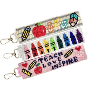 May include: Three beaded wristlet keychains with silver clasps. The keychains feature school-themed designs, including pencils, apples, crayons, and the words "TEACH LOVE INSPIRE." The keychains are in white, pink, and gray.