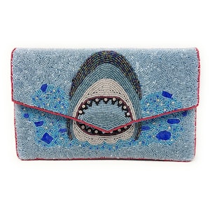 May include: A beaded clutch featuring a shark design. The clutch is light blue with a red trim and a shark's open mouth in the center. The shark is made of white, black, and blue beads, with blue water details.