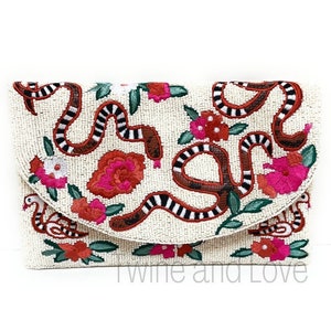 May include: A white beaded clutch with a colorful embroidered design of snakes, flowers, and birds. The design is in shades of red, pink, orange, black, and green.