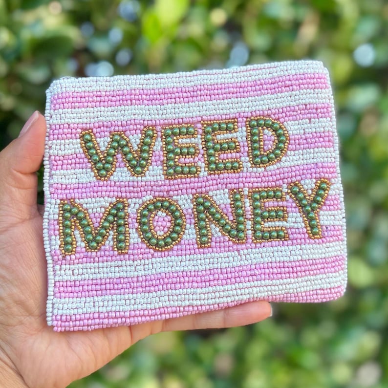 May include: A beaded pouch with pink and white stripes. The words "WEED MONEY" are spelled out in green and gold beads. The pouch is rectangular and appears to be held by a person.