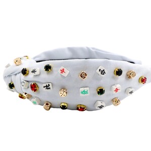 A white headband with a twisted design, adorned with various colorful gemstone studs and Mahjong tile charms, conveying a playful and eclectic style.