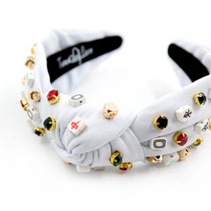 A white headband with a twisted design, adorned with various colorful gemstone studs and Mahjong tile charms, conveying a playful and eclectic style.