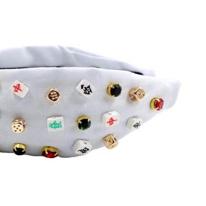 A white headband with a twisted design, adorned with various colorful gemstone studs and Mahjong tile charms, conveying a playful and eclectic style.