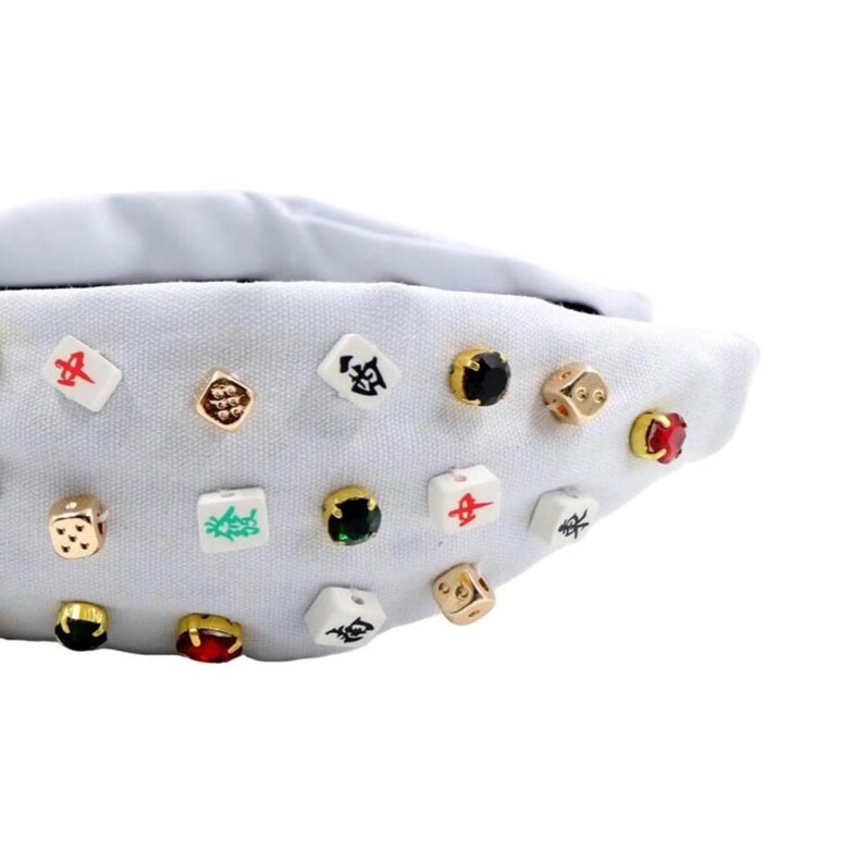 A white headband with a twisted design, adorned with various colorful gemstone studs and Mahjong tile charms, conveying a playful and eclectic style.