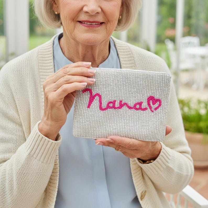 May include: A beaded silver clutch with the word "Nana" embroidered in pink script, accompanied by a pink heart. The clutch is rectangular and held by a person wearing a cream-colored cardigan.