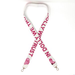 Pu&ograve; includere: Tracolla per borsa in perle bianche con testo ricamato rosa che recita "In Dolly We Trust" e un'immagine rosa di un viso di donna in stile cartone animato su ogni estremit&agrave;.