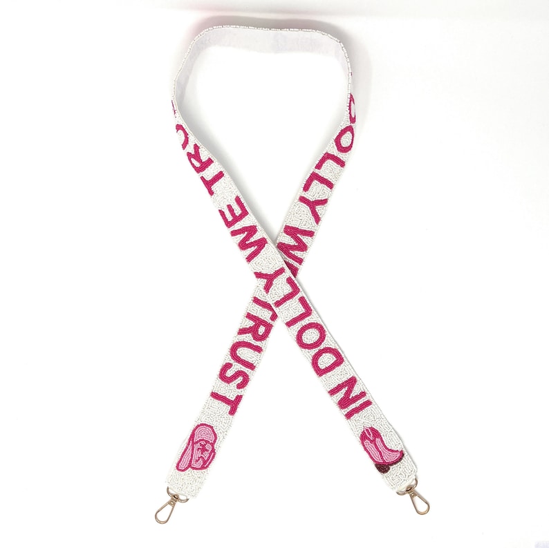 Pu&ograve; includere: Tracolla per borsa in perle bianche con testo ricamato rosa che recita "In Dolly We Trust" e un'immagine rosa di un viso di donna in stile cartone animato su ogni estremit&agrave;.