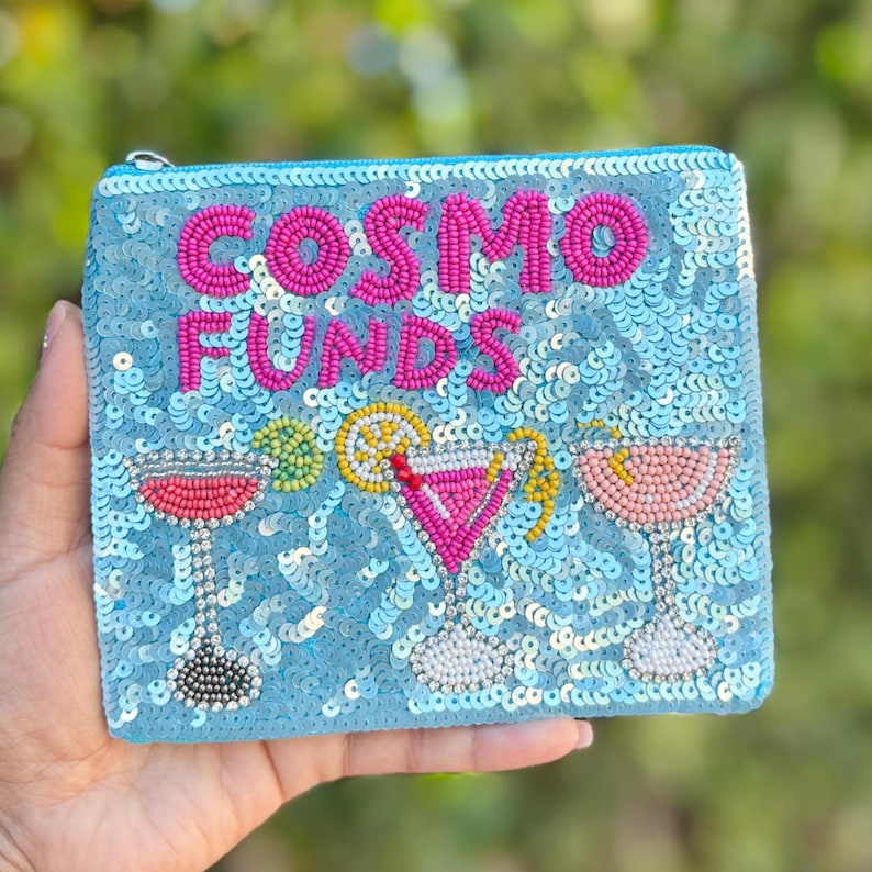 May include: A light blue sequined cosmetic bag with the words "COSMO FUNDS" in pink beaded letters. The bag features beaded cocktail designs in pink, yellow, and green. A zipper closure is visible.