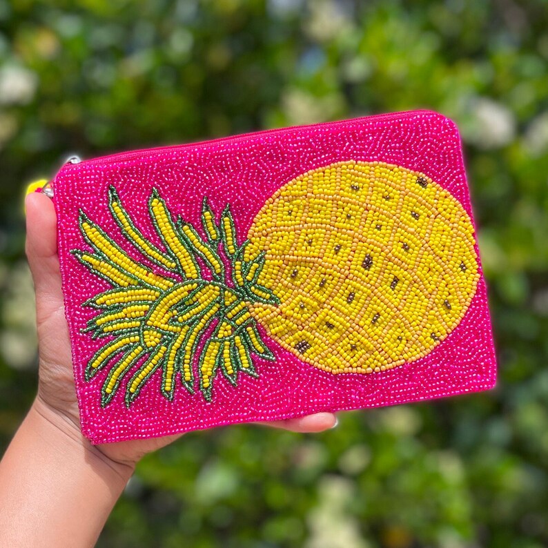 May include: A pink beaded clutch with a yellow pineapple design. The pineapple is made of yellow and orange beads, and the leaves are green.