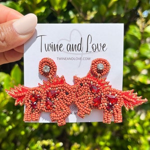 May include: A pair of red beaded earrings shaped like a unicorn with a rhinestone on each ear. The earrings are on a white card with the text "Twine and Love" and "twineandlove.com".