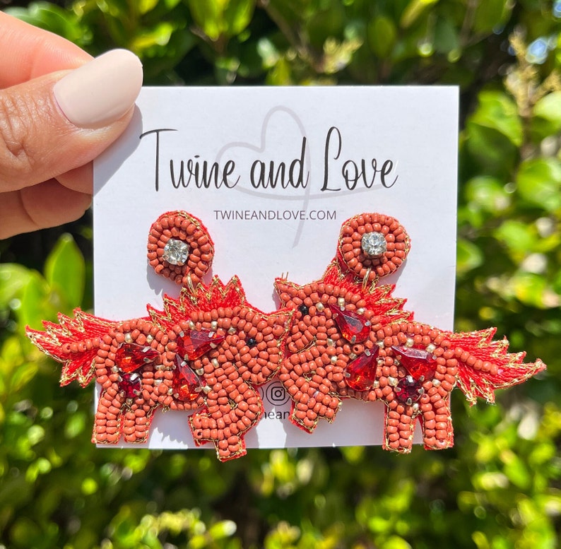May include: A pair of red beaded earrings shaped like a unicorn with a rhinestone on each ear. The earrings are on a white card with the text "Twine and Love" and "twineandlove.com".