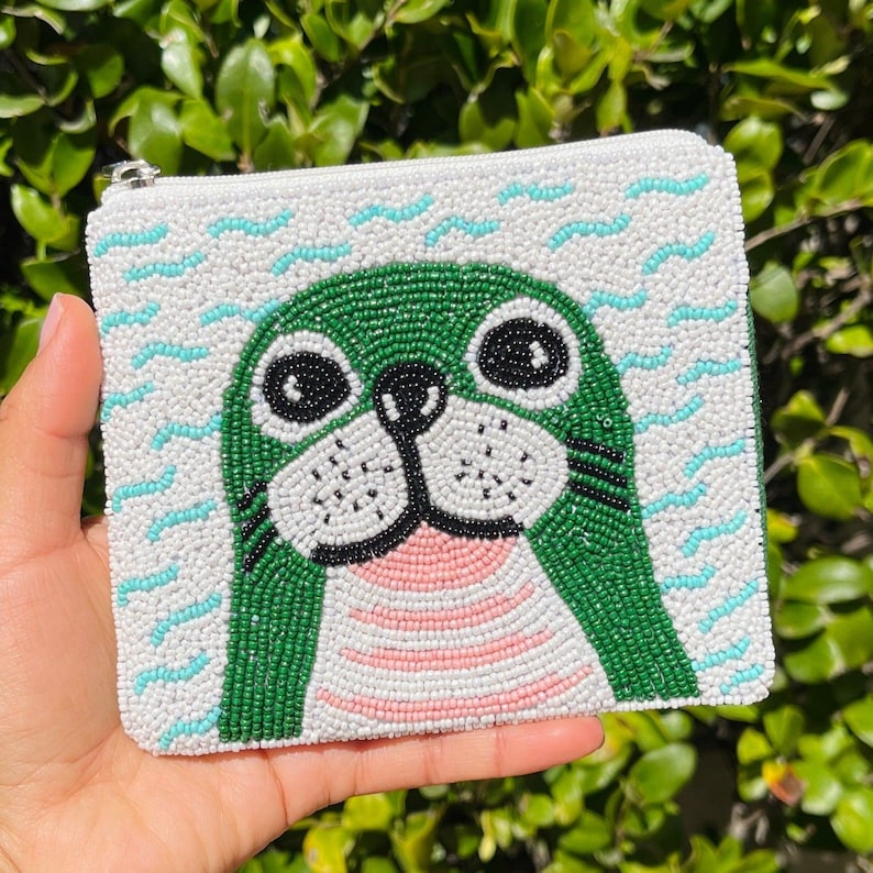 K&ouml;nnte beinhalten: Eine wei&szlig;e, perlenbesetzte Tasche mit Rei&szlig;verschluss und einem gr&uuml;nen Seehund-Design. Der Seehund hat schwarze Augen, Schnurrhaare und einen rosa Mund. Der Hintergrund hat wei&szlig;e Perlen mit blauen Wellenmustern.